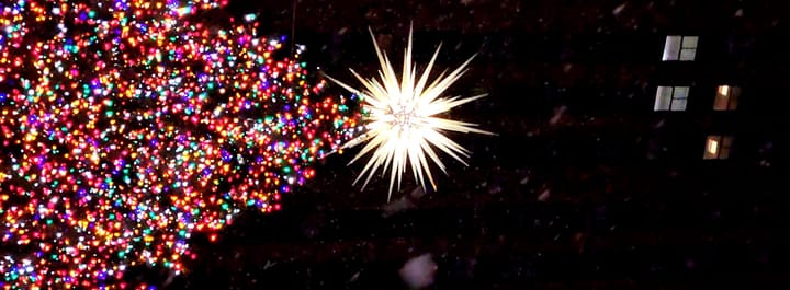 Colorful Christmas tree lights, the spiky white star on top, snow flakes blurry in the air, lit windows several stories above.